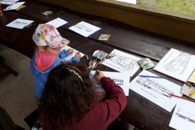 Dziecko siedzi przy drewnianym stole i koloruje obrazki. Na stole rozłożone są kredki i kartki z rysunkami. Dziecko ma na głowie kolorową opaskę z papieru.