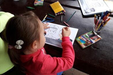 Młoda dziewczynka w różowym swetrze koncentruje się na kolorowaniu obrazka przedstawiającego postać ludzką. Na stole leżą kredki, flamastry i ołówki. W tle widoczne są inne materiały plastyczne oraz kolorowanka.