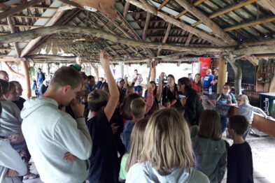 Grupa ludzi zgromadziła się w drewnianym przybytku o otwartej konstrukcji, słuchając osoby stojącej pośrodku. Większość uczestników skupia uwagę na mówcy, trzymając w dłoniach aparaty fotograficzne i smartfony. Konstrukcja, pod którą się znajdują, jest wykonana z drewnianych bali i pokryta strzechą.