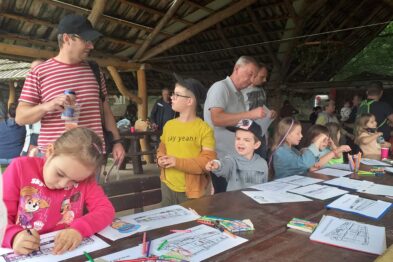 Grupa dzieci siedzi przy długich stołach, skupiona na kolorowaniu i rysowaniu obrazków. Dorosłe osoby chodzą wokół, obserwując i pomagając młodszym uczestnikom. Wszyscy znajdują się pod zadaszeniem w otoczeniu zieleni, co wskazuje na zajęcia na świeżym powietrzu.