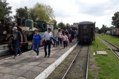 Ludzie wysiadają z historycznego pociągu na muzealnej stacji kolejowej. Po prawej stronie widać wagon, a po lewej parowóz z wieloma rurami i elementami na zewnątrz. Tło zdobią drzewa i niebiesko-białe niebo z chmurami.