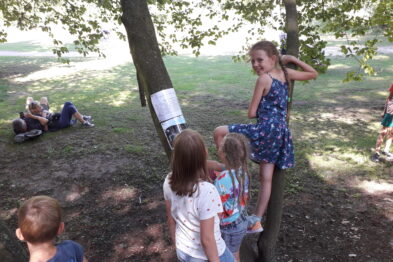 Grupa dzieci bawi się na zewnątrz w cieniu drzew. Jedno z nich wspina się na drzewo, trzymając się gałęzi, podczas gdy inne obserwują. W tle dwójka dzieci odpoczywa na ziemi kładąc się na trawie.