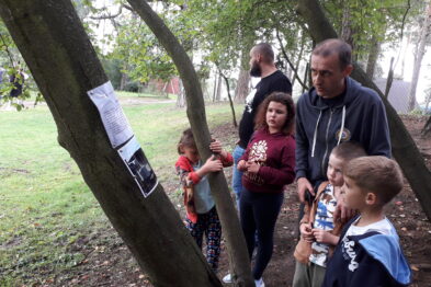 Grupa osób, w tym dzieci i dorośli, stoi w otoczeniu drzew. Na jednym z drzew wisi kilka kartek papieru, które uczestnicy obserwują. Dzieci trzymają w rękach kredki do rysowania.