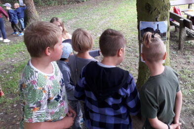 Grupa dzieci stoi w lesie, zwrócona twarzami do drzewa, na którym wisi kolorowa fotografia. Na pierwszym planie widać czterech chłopców obserwujących zdjęcie; jeden z nich ma na głowie zieloną czapkę. W tle rozpoznajemy teren zielony, z ławkami i innymi uczestnikami wydarzenia rozmytymi w oddali.