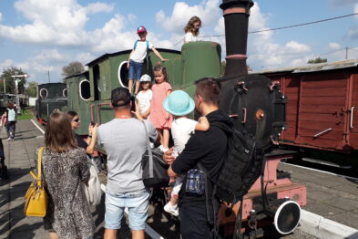 Grupa osób, w tym dzieci i dorośli, znajduje się wokół starej lokomotywy parowej na stacji kolejowej. Dzieci wchodzą na parowóz, pod czujnym okiem dorosłych, co wskazuje na interaktywny charakter wystawy. W tle widać także inne wagony kolejowe, co świadczy o bogatej ekspozycji muzealnej.