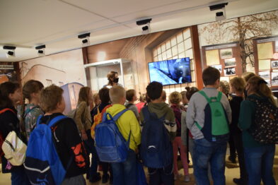Grupa dzieci z plecakami ogląda prezentację w muzeum kolejnictwa; stoi skupiona przed ekranem, na którym wyświetlane są treści związane z kolejami. Wnętrze jest jasne, a na ścianach widoczne są różne eksponaty. Przewodnik muzealny w mundurze stoi po prawej stronie, towarzysząc dzieciom w ich edukacyjnej wizycie.