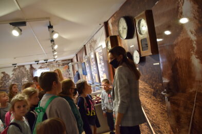 Grupa dzieci z przewodnikiem zwiedza ekspozycję muzealną związaną z kolejnictwem. W tle widoczne są wystawione eksponaty, takie jak zegary i fotografie kolejnictwa. Dzieci słuchają opowieści, niektóre z nich mają plecaki.
