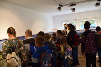 Grupa dzieci stoi przed ekspozycją w muzeum, skupiona na słuchaniu osoby prowadzącej. Dzieci w różnorodnych ubraniach, niektóre z plecakami, patrzą w kierunku prezentowanych eksponatów. W tle widać ścianę z fotografiami oraz modele kolejek, na które uczestnicy lekcji zwracają uwagę.