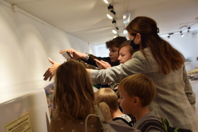 Grupa dzieci wraz z opiekunką w muzeum uczestniczy w lekcji edukacyjnej. Stoją wokół ekspozycji, gdzie dzieci wskazują palcami na fotografie i modele kolejowe. Opiekunka wydaje się tłumaczyć i objaśniać treści związane z historią kolei.