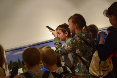 Grupa dzieci stoi skoncentrowana przed przeszkloną witryną z eksponatami kolejowymi; jedno z nich wskazuje palcem na jakiś obiekt. Osoba dorosła, stojąca obok, podtrzymuje wzniesioną dłoń dziecka, wspierając jego zainteresowanie. Dookoła rozświetloną salę wypełniają barwne obrazy oraz modele kolejowe prezentujące różne aspekty tematyczne związane z koleją.