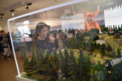 Grupa osób ogląda dużą makieta kolejową umieszczoną pod osłoną szklanej witryny. Makieta pokazuje szczegółowy krajobraz z torami kolejowymi, miniaturkowymi drzewami i budynkami. Odbicie jednej z osób jest widoczne na szybie witryny.