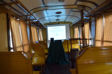 Wewnątrz drewnianego wagonu kolejowego ustawiono projektor oraz ekran, na którym wyświetlana jest prezentacja. Drewniane ławki pasażerskie rozmieszczone są po obu stronach wagonu. Zadbane wnętrze sugeruje przygotowanie do zajęć edukacyjnych związanych z historią kolei.