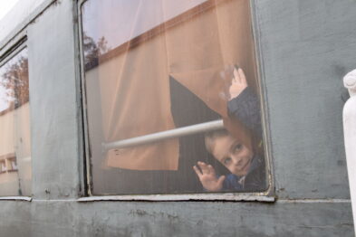 Dziecko macha ręką przez okno pociągu. Okno jest częściowo zasłonięte przez brązową zasłonę. W tle widać szare ściany budynku.