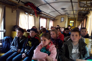 Grupa dzieci siedzi na drewnianych ławkach wewnątrz wagonu kolejowego, zwrócona w stronę kamery. Wagon wyposażony jest w metalowe bagażniki i okna z żółtymi zasłonkami. Dzieci wydają się być zaangażowane w lekcję lub prezentację.