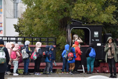 Grupa dzieci stoi na platformie kolejowej przy wagonie, słuchając przewodnika. Dzieci mają na sobie kolorowe ubrania i plecaki, a niektóre z nich noszą czapeczki. Przewodnik, ubrany w ciepłą kurtkę, gestykuluje i opowiada o wagonie.