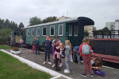 Grupa dzieci stoi obok zielonego wagonu kolejowego, który wydaje się być częścią ekspozycji muzealnej. Dzieci są ubrane w różnorodne ubrania i wydają się posiadać plecaki. W tle widoczna jest również ciemniejsza lokomotywa oraz budynki, które mogą być częścią kompleksu muzealnego.