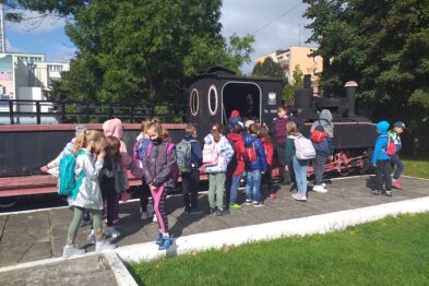 Grupa dzieci w wieku szkolnym jest zgromadzona wokół czarnej lokomotywy parowej na zewnątrz. Uczestnicy noszą różnorodną odzież sportową i plecaki. Lokomotywa jest umieszczona na wystawie, a dzieci wydają się być zainteresowane eksponatem.