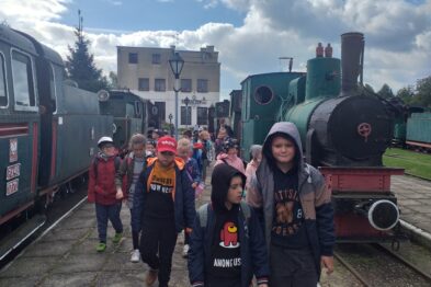Grupa dzieci w wieku przedszkolnym i wczesnoszkolnym spaceruje między starymi lokomotywami na terenie muzeum kolejowego. Dzieci są ubrane w kurtki i czapeczki, kilka z nich nosi plecaki. W tle widać zabytkowe parowozy, które są wystawione na zewnątrz.