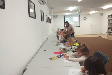 Grupa dzieci siedzi przy długim stole, skupiona na kolorowaniu lub pisaniu. Dorośli prowadzą zajęcia, jeden z nich stać przy tablicy, a drugi asystuje przy stole. Sala lekcyjna jest jasna i wyposażona w półki z książkami oraz tablicę.