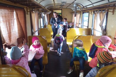 Grupa dzieci w towarzystwie dorosłej osoby znajduje się wewnątrz wagonu kolejowego o drewnianych ławkach i żółtym wnętrzu. Dzieci mają na sobie kolorowe czapki i warstwowe ubrania, niektóre siedzą, inne stoją lub poruszają się wzdłuż przejścia. Wagon jest oświetlony naturalnym światłem, a okna przyozdobione są zasłonami.