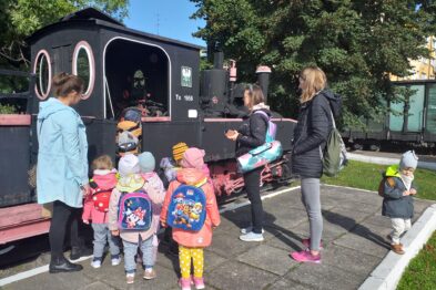 Grupa dzieci stoi przed czarną lokomotywą wąskotorową, posłuchując opowieści przewodniczki. Przewodniczka, trzymająca materiały edukacyjne, wskazuje na detal maszyny. Dzieci są ubrane w kolorowe plecaki i odwrócone twarzami w stronę lokomotywy, co świadczy o zainteresowaniu tematyką kolejnictwa.