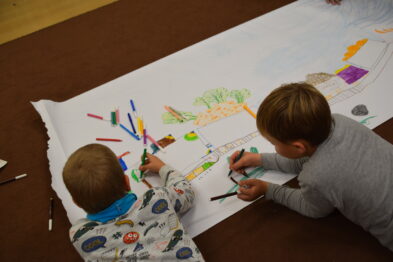 Dwoje dzieci siedzi na podłodze i wspólnie rysuje kolorowymi kredkami na dużym arkuszu papieru, który przedstawia tor kolejowy i otoczenie. Ubrani są w ciepłe, jasne ubrania, a wokół nich rozrzucone są kolorowe pisaki i kredki. Całość rozgrywa się w jasnym pomieszczeniu z widocznym dywanem i meblami.