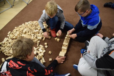 Grupa dzieci siedzi na podłodze i bawi się drewnianymi klockami. Budują z nich struktury przypominające tory kolejowe. Wokół nich rozrzucone są inne klocki i elementy zestawu.