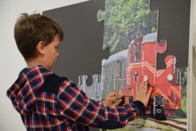 Chłopiec w kratowanej koszuli układa duże puzzle przedstawiające czerwoną lokomotywę. Koncentruje się na dopasowywaniu kawałków, pracując przy ściennej planszy. Zajęcie to jest częścią edukacyjnego programu poświęconego historii i technologii kolejnictwa.