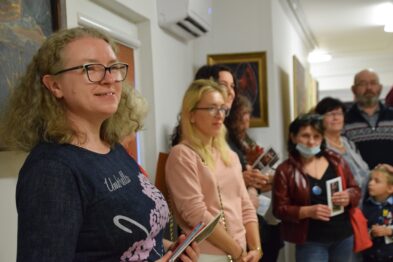 Grupa osób stoi wewnątrz galerii, skupiając swoją uwagę na czymś poza kadrem zdjęcia. Z przodu po lewej stronie widać uśmiechniętą kobietę trzymającą kartkę lub broszurkę. Na ścianach za postaciami eksponowane są obrazy w ramach, a uczestnicy wydarzenia wydają się być zainteresowani wystawą.