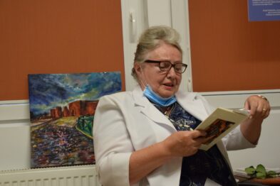 Kobieta w białym żakiecie czyta z otwartej książki. Na ścianie za nią wisi kolorowy obraz przedstawiający scenę kolejową. Pani ma na sobie niebieską apaszkę i okulary.
