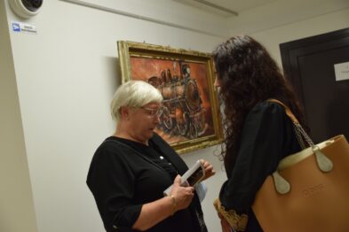 Dwie kobiety stoją naprzeciwko siebie i rozmawiają w galerii, jedna z nich trzyma telefon komórkowy w dłoniach. W tle widoczne jest zawieszone na ścianie obrazu o tematyce kolejowej, przedstawiające lokomotywę i postacie. Ściany galerii są jasne, a oświetlenie skupia się na dziełach sztuki.