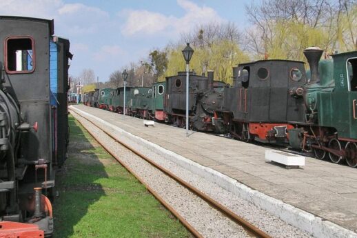 Szlak kolejowy wiodący przez muzeum otaczają z obu stron stare lokomotywy i wagony kolejowe w różnych kolorach ustawione równolegle względem siebie. Tor kolejowy wyposażony jest w drewniane podkłady, a wzdłuż toru znajdują się metalowe latarnie. Tło obrazu stanowi bezchmurne niebo i rozproszone drzewa w tle.