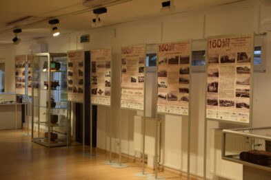W muzeum ustawiono panele wystawiennicze prezentujące fotografie i opisy historyczne, które są zawieszone na ścianach i wsparte na metalowych konstrukcjach. Na przedniej części zdjęcia widać szklane witryny z eksponatami oraz modele pociągów. Oświetlenie podkreśla prezentowane treści i tworzy przyjemną atmosferę dla zwiedzających.