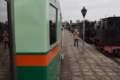 Skład kolejowy z wagonem motorowym o zielono-pomarańczowym malowaniu stoi na peronie; w tle widoczna jest parowa lokomotywa i grupa ludzi. Peron pokryty kostką brukową, jest pochmurny dzień. Osoby na peronie ubrane są w ciepłe kurtki, sugerując, że jest to chłodniejsza pora roku.