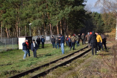 Grupa ludzi stoi obok torów kolejowych, wśród nich niektórzy fotografują wydarzenie. Osoby są ubrane w ciepłe kurtki, co wskazuje na chłodną pogodę. W tle widać drzewa i niewielką budowlę przy torach.