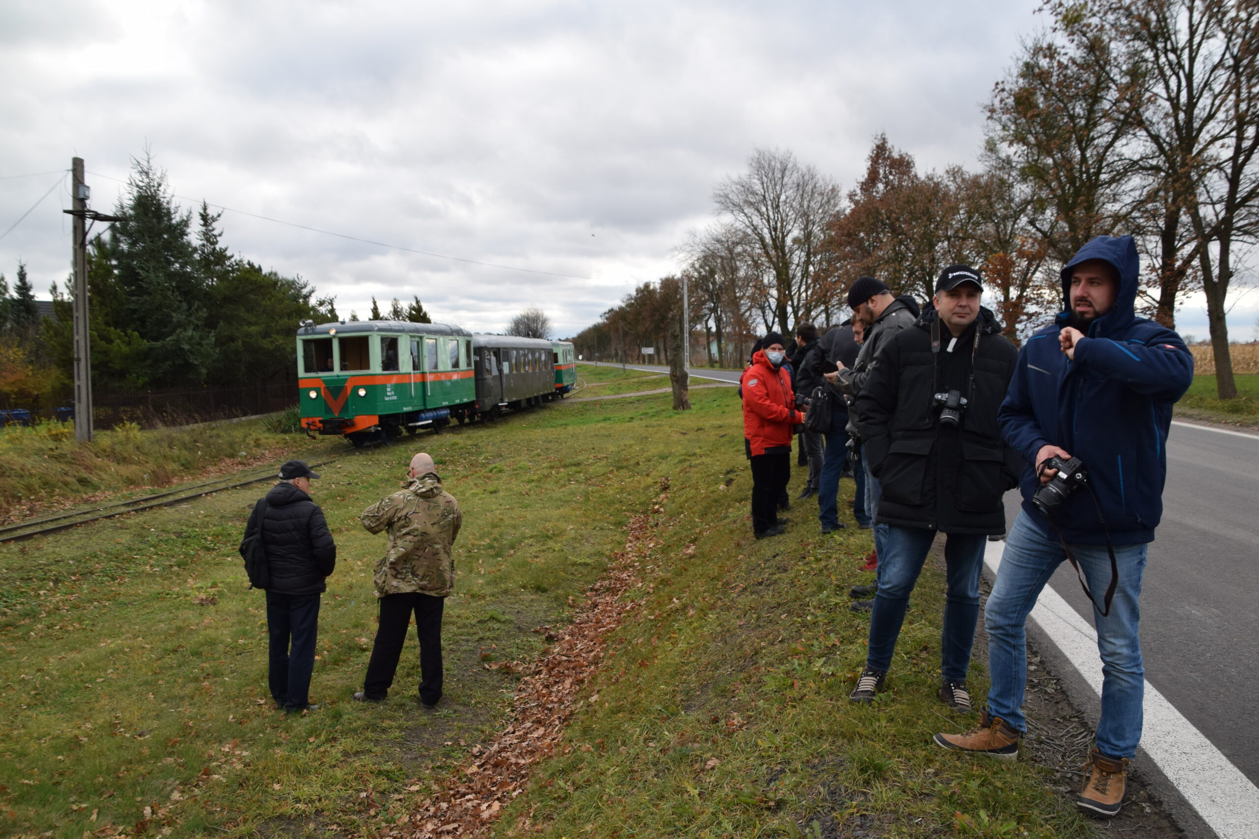 Grupa osób stoi na trawiastym terenie obok torów kolejowych, skierowani są w stronę nadjeżdżającego pociągu. Pociąg składa się z zielonych wagonów, które są charakterystyczne dla kolei wąskotorowej. Niektórzy z obecnych mają w rękach aparaty fotograficzne, prawdopodobnie aby uwiecznić wydarzenie.