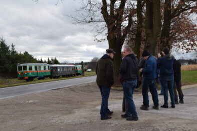 Grupa osób stoi przy drodze w oczekiwaniu na zbliżający się pociąg. Skład kolejowy, składający się z dwóch wagonów motorowych i wagonu osobowego, jest widoczny w tle. Drzewa po prawej stronie dodają urok krajobrazowi.