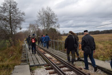 Grupa osób spaceruje po drewnianym mostku kolejowym nad rzeką, otoczeni przez pola i niewielkie zadrzewienia. Niektórzy uczestnicy mają plecaki i wydają się być w średnim wieku, ubrani są w ciepłe kurtki i czapki. Tor kolejowy na mostku składa się z dwóch szyn i zamocowanych poprzecznie belek drewnianych.