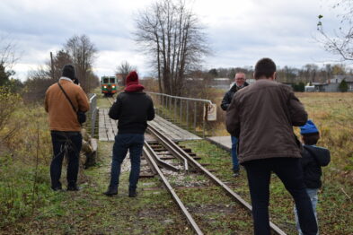 Grupa osób stoi na nasypie kolejowym i ogląda nadjeżdżający pociąg. Osoby mają na sobie ciepłe ubrania i niektóre z nich robią zdjęcia. Tor kolejowy wiedzie przez teren z drzewami, a w tle widać nadjeżdżający skład kolejowy.
