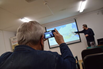Starszy mężczyzna robi zdjęcie telefonem komórkowym; celuje w ekran, na którym wyświetlana jest prezentacja z mapą. W tle widać prelegenta wskazującego na wyświetlane treści. Pomieszczenie wygląda na salę wykładową z rzędami krzeseł skierowanymi w stronę ekranu.