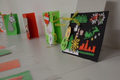 Ręcznie wykonane kartki świąteczne stoją wyeksponowane na białym stole. Każda z kartek jest ozdobiona kolorowymi wycinankami, naklejkami i elementami charakterystycznymi dla okresu świątecznego. Dominują motywy kolejowe, Mikołaje i choinki, które nawiązują do tematyki kolei i nadchodzących świąt.