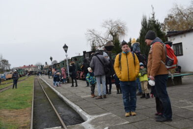 Ludzie zebrali się na peronie przy torach wąskotorowych, czekając na wydarzenie. Dzieci i dorośli w zimowych ubraniach rozmawiają i spędzają czas na zewnątrz. W tle widać stojący pociąg oraz osoby przechadzające się między kolejowymi pojazdami.