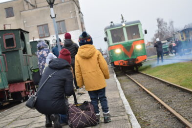Grupa ludzi oczekuje na zielony wąskotorowy pociąg zbliżający się po torach; wśród nich są dzieci i dorośli ubrani w zimowe kurtki. Niebo jest zachmurzone, co wskazuje na zimową aurę. W tle widoczne są zabudowania i wagony kolejowe, co podkreśla kolejowy charakter wydarzenia.