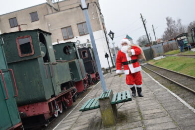 Święty Mikołaj w czerwonym stroju trzyma w rękach dzwonek i maszeruje wzdłuż torów kolejowych obok zielonej lokomotywy wąskotorowej. Na peronie widać drewnianą ławkę, a w tle budynek stacji. Torowisko jest mokre, co sugeruje niedawne opady deszczu.