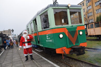 Strojny w tradycyjny czerwony kostium Świętego Mikołaja z białym zarostem stoi obok zielonego wagonu kolejowego z pomarańczowymi elementami i naprzemiennym pasem w kształcie litery 