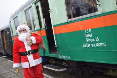 Osoba przebrana za Świętego Mikołaja stoi obok zielonego wagonu kolejowego, trzymając dzwonek w lewej dłoni i worek na prezentach w prawej. Na boku wagonu widnieją białe napisy z informacjami o czasie odjazdu, ilości miejsc i numerem bocznym. Pogoda jest pochmurna, co wskazuje na zimowy charakter wydarzenia.