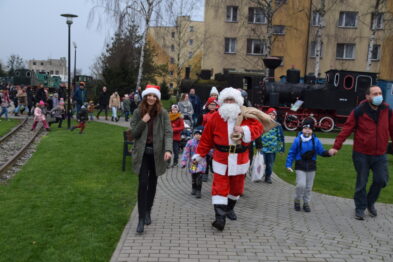 Grupa ludzi, wśród których widać osobę przebraną za Świętego Mikołaja, idzie chodnikiem obok torów kolejowych. W tle znajduje się parowóz oraz kilka budynków. Osoby na zdjęciu są ubrane w zimowe odzieże, a dzieci trzymają w rękach balony.