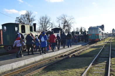 Grupa osób w różnym wieku stoi na peronie obok pociągu o zielonej barwie na wąskotorowej stacji. Ludzie są ubrani w zimowe kurtki, niektórzy mają na głowach karnawałowe nakrycia. Peron i tory biegną równolegle, z tłumem ludzi zbliżającym się do pociągu ciągniętego przez lokomotywę.
