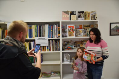 Mężczyzna robi zdjęcie smartfonem dwóm osobom stojącym obok półki z książkami. Kobieta i dziecko trzymają w rękach kolorowy model pociągu. W tle widoczne są regały z różnymi przedmiotami i książkami.