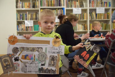 Chłopiec trzyma w ręku kolorową, ręcznie wykonaną pracę plastyczną, na której widnieją obrazki i elementy związane z tematyką kolejnictwa. Siedzi przy stole w pomieszczeniu, które przypomina bibliotekę, w tle widać półki z książkami. Inne dzieci również pracują przy stołach, a dorośli ich asystują.